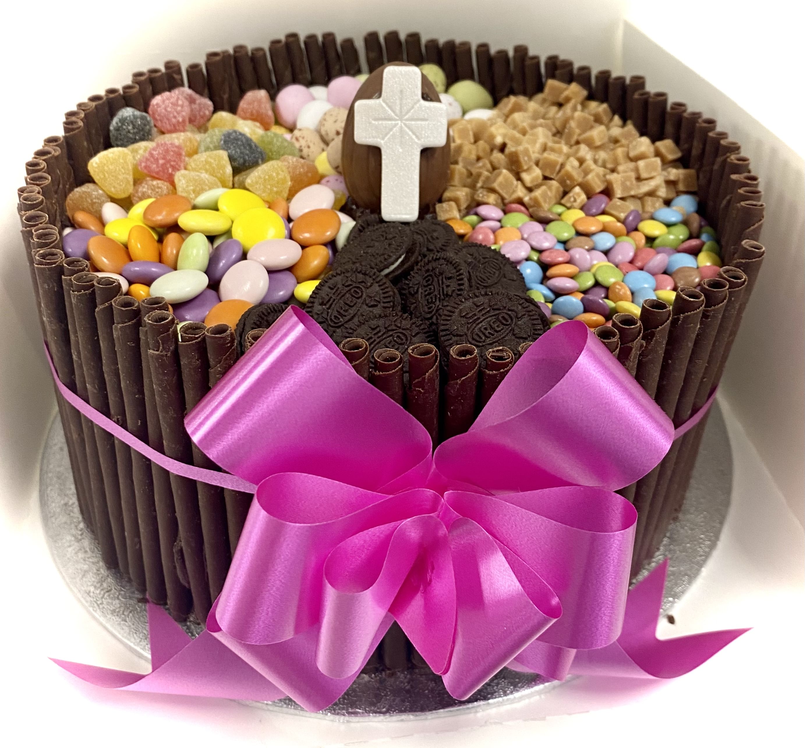 A chocoholic's dream, this chocolate layer cake is decorated with chocolate sticks around the sides and features sections filled with colorful candies, cookies, and fudge on top. A white cross stands in the center, while a pink ribbon with a large bow is tied around the middle of the cake.